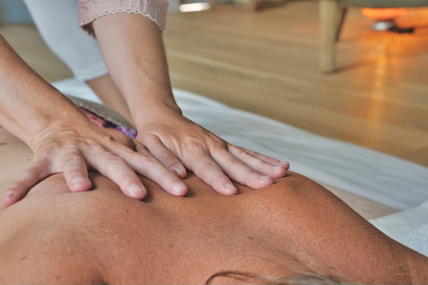 A close-up of two hands giving a back massage to a person lying face-down on a white surface in a warmly lit room with wooden flooring.