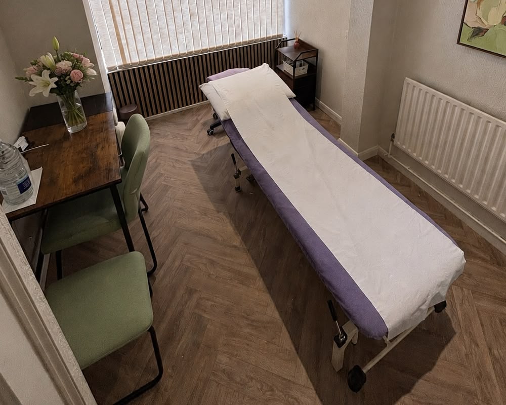 A treatment room with a padded massage table covered in paper, a wooden desk with a vase of flowers, two green chairs, and a window with vertical blinds letting in natural light.